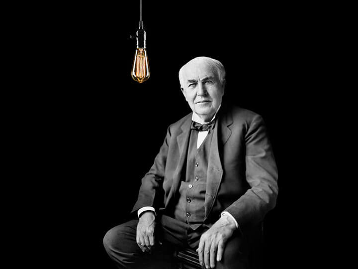 Thomas Edison sitting with an Edison lightbulb hanging next to him with a black background